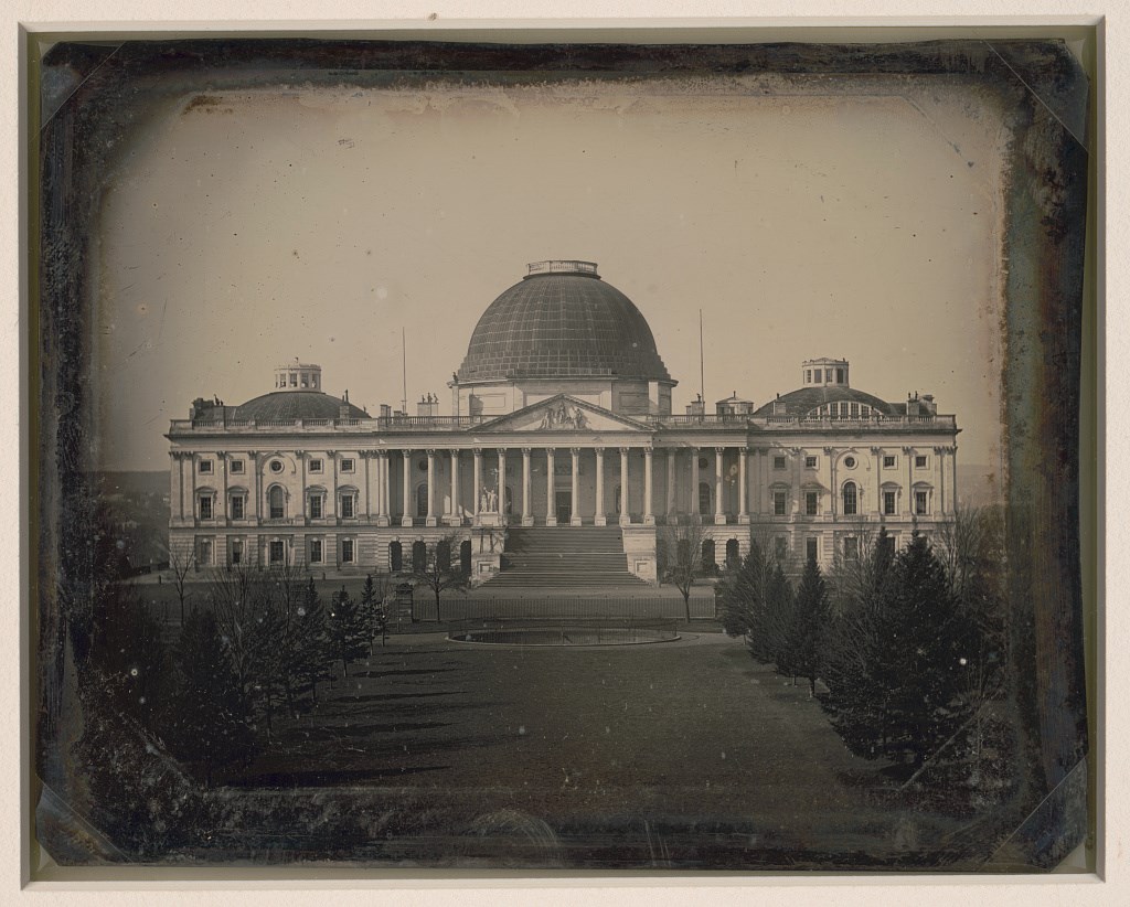 Black and white photo of US Capitol building in 1846. Building is 3 stories, long, and white with 3 dark domes, the middle dome is the largest. Grand stairs lead to pillars in front of main door.
