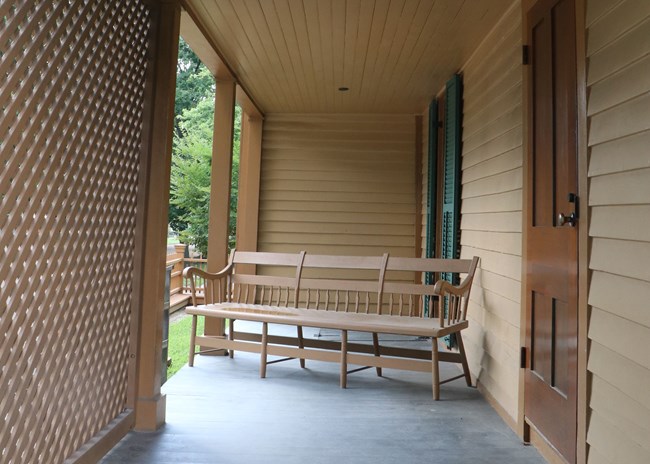 covered porch with light blue wooden floor, wooden coffee colored screen on left, and door on the wall on right. Bench sits on porch, next to window with dark green shutters