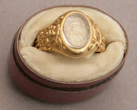 a gold ring with a clear glass case, a thin brown piece of hair is curled inside the case