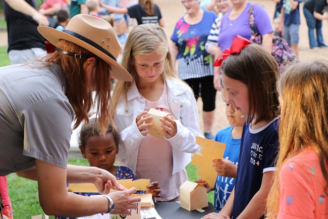 A park ranger leans over a table while helping several young girls with a craft activity.