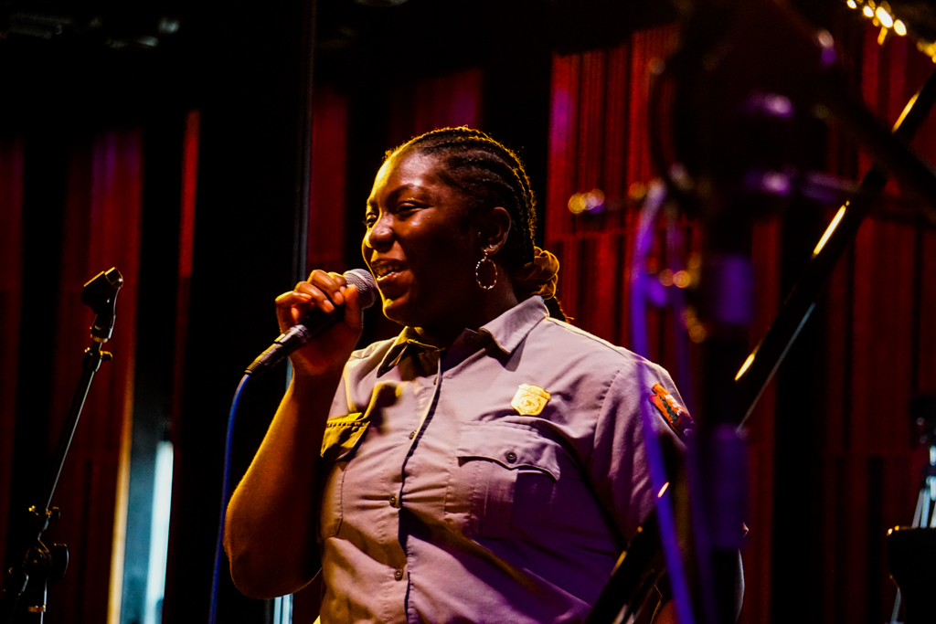 a park ranger in uniform holds a microphone against a red curtain background