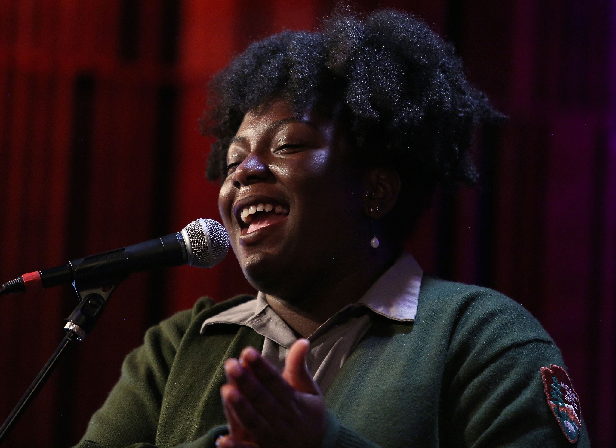 A uniformed park ranger sings and claps in front of a dark red curtain background.