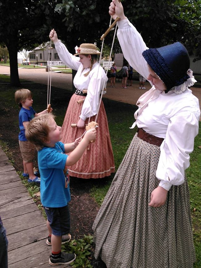 Living History volunteers in 1860s period clothing demonstrate how to play with old puppet toys.