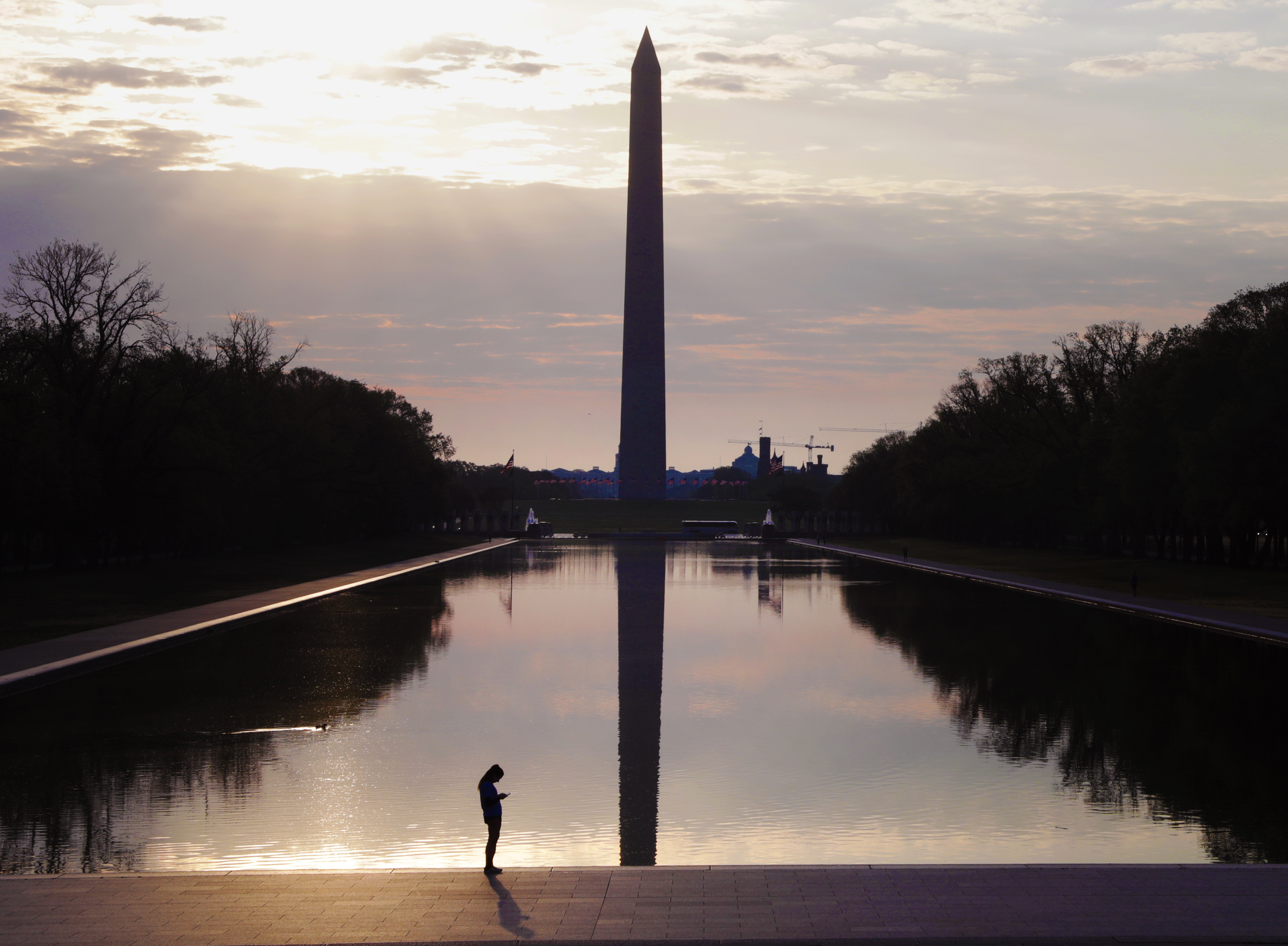 The Washington Monument stands silhouetted against the rising sun, reflecting in the still Reflecting Pool.