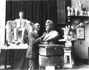 Man in art studio working on bust of statue with statue of man sitting in chair behind him