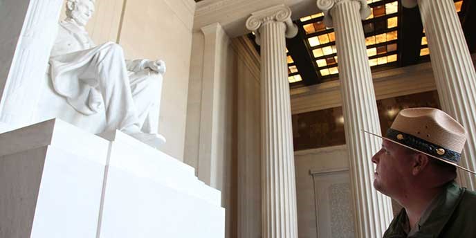 A man with hat gazes at a white statue sitting in chair behind him are columns