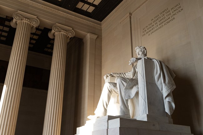 A ray of sunlight illuminates the marble statue of Abraham Lincoln in the Lincoln Memorial. Tall ionic columns appear on the left of the image.