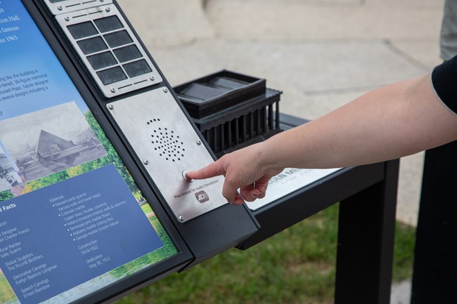 A visitor presses the audio description button on an information panel with bronze model of the Lincoln Memorial.