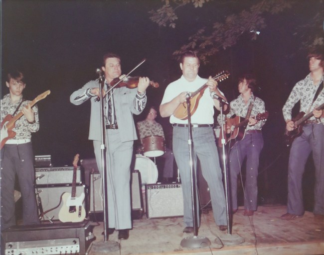 A group of six men on a stage playing guitars, drums, a fiddle, and a mandolin.