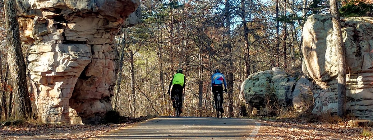 Two road bikers riding by a rock formation on a paved highway