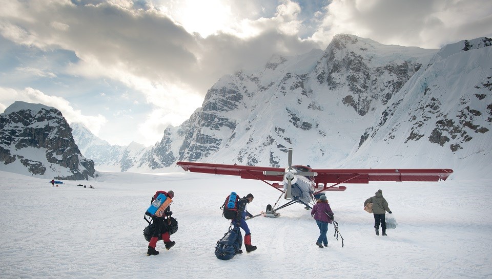 A park commercial operator picks visitors up with a plane on skis on a snowy mountain. Four hikers on a snowy mountain walk with gear to a plane on skis.