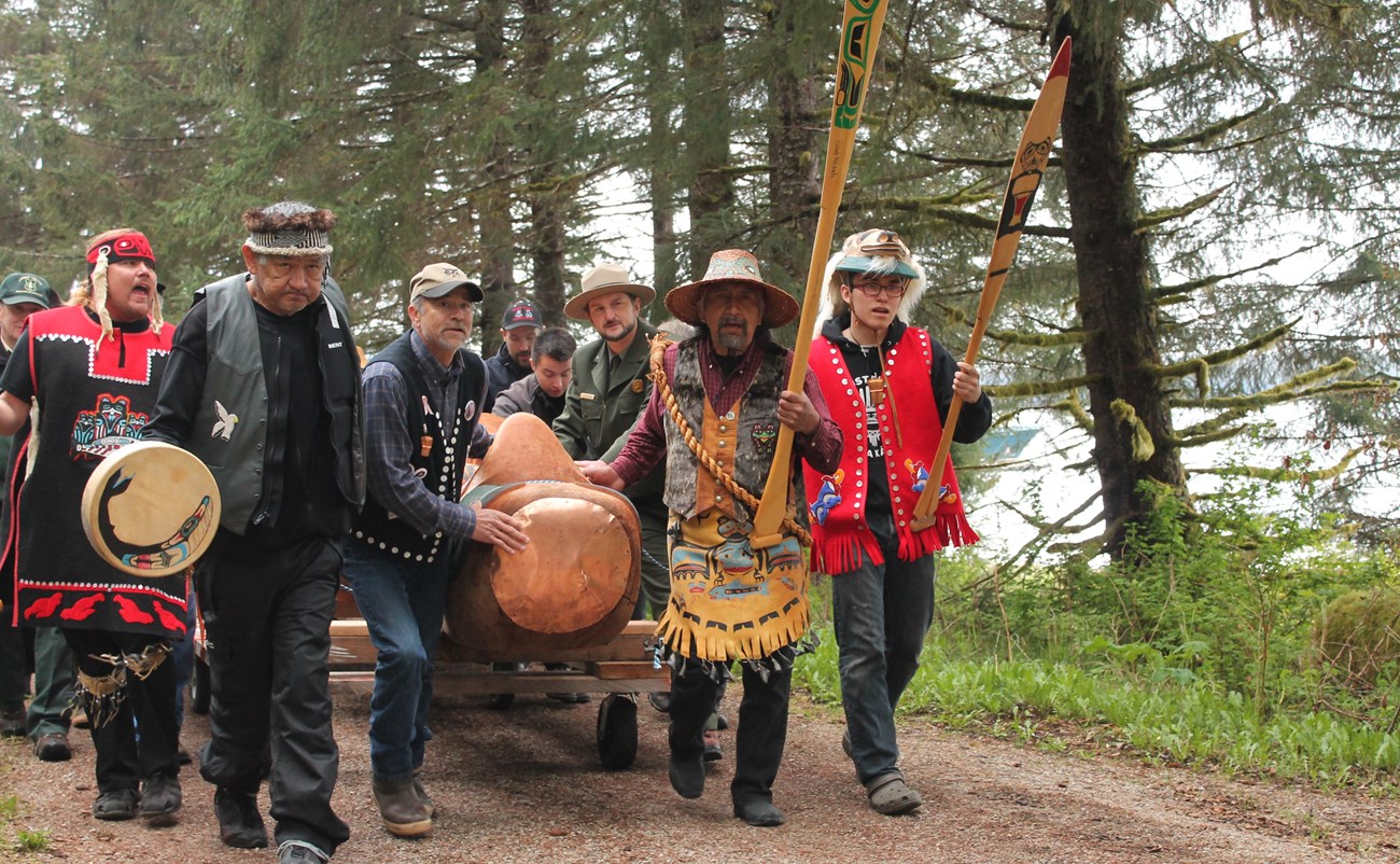 A group of men carry a totem pole. They are in ceremonial dress.