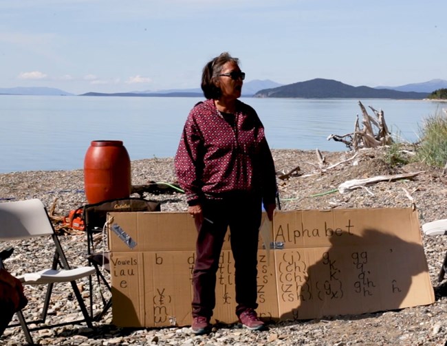 A female elder stands in front of a cardboard sign that says "Dena'ina Alphabet"