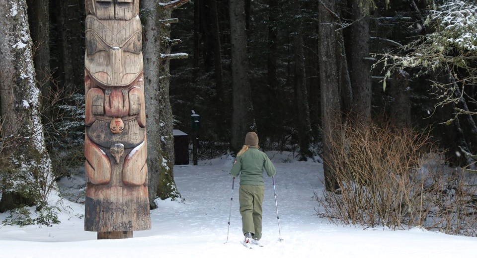 A female park ranger skis next to a totem pole on a snowy trail into the woods