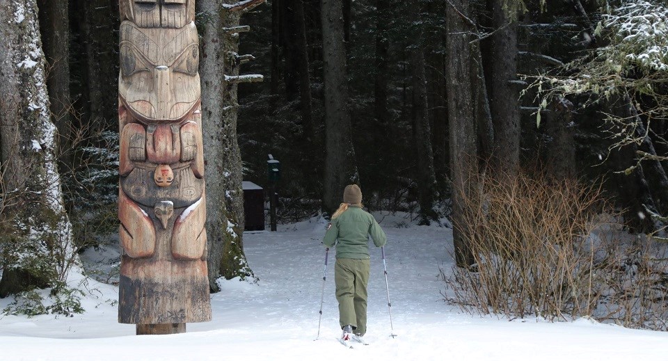 Skiing in Sitka A female park ranger skis next to a totem pole on a snowy trail into the woods