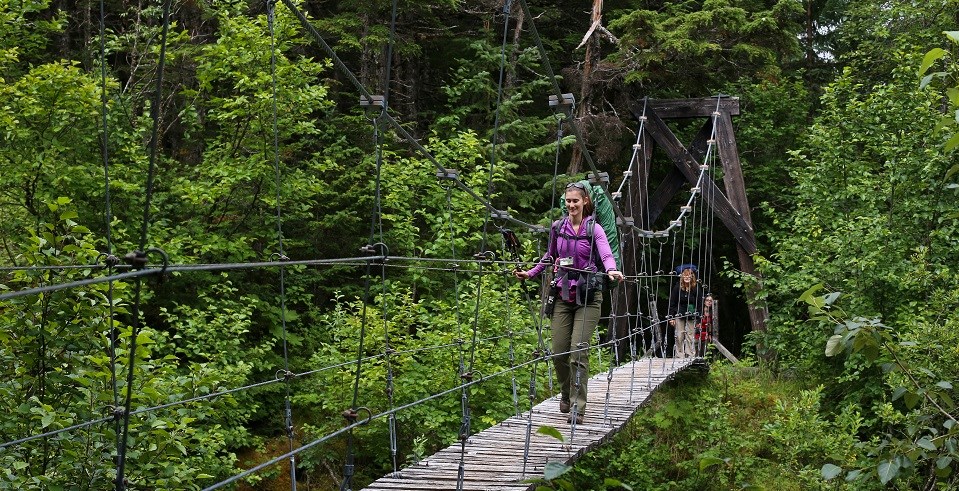 A woman with a backpack walks across a suspension bridge in a wooded area