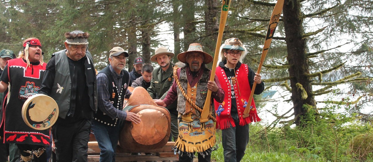 A group of male individuals are walking and singing while carrying a totem pole. Some of the tribal members are wearing tribal hats and vests. The primary colors are black and red, although there is one person wearing a tan colored vest.