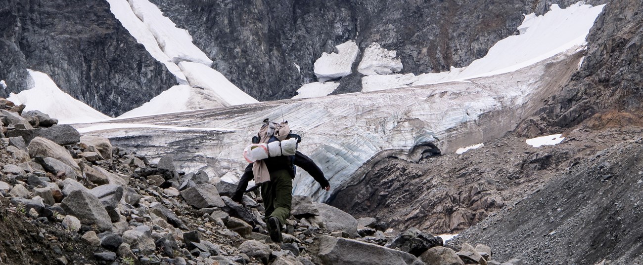 a person with a large backpack walks up a rocky ravine in front of a small glacier