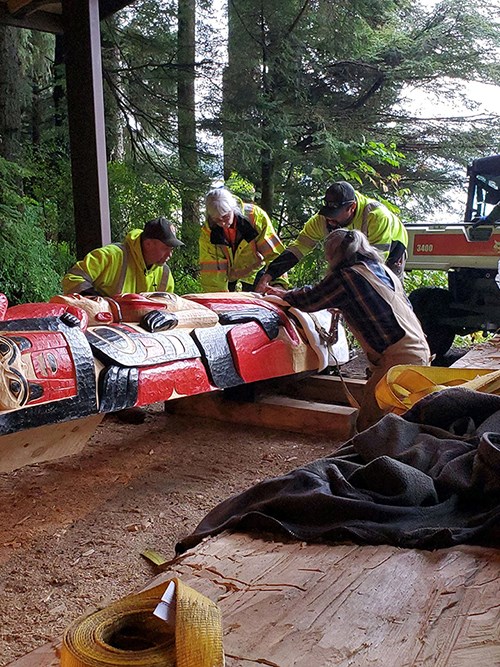 Four people prepare a totem pole to move. The pole is resting horizontally on wood blocks.