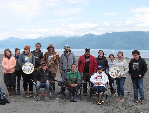 A group of Alaska Natives pose on the beach. Three are holding native crafts including two drums.