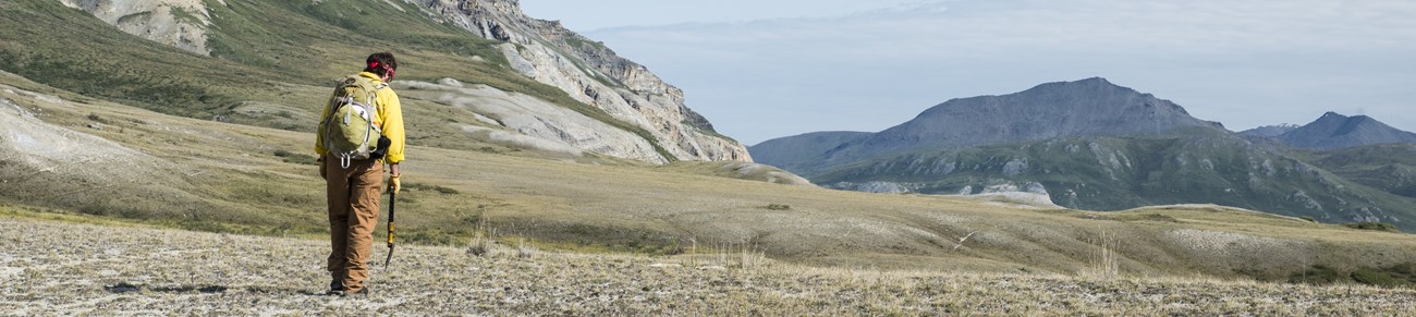 a man wearing a yellow jacket and green backpack holds a shovel as large mountains extend out toward the horizon.