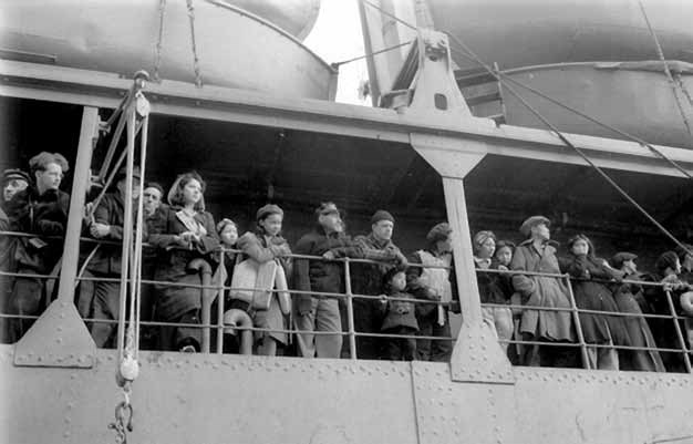 Pribilof villagers, here lining the railing of the USAT Delarof on the day of their departure (June 15, 1942), were evacuated with only a few hours notice and no idea of their ultimate destination. A group of warmly-dressed people stand on a ship balcony looking over the rail with expressions of uncertainty.
