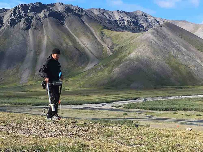 A man walks the landscape holding a tool, the tool is facing the ground. A man walks the landscape holding a tool, the tool is facing the ground.