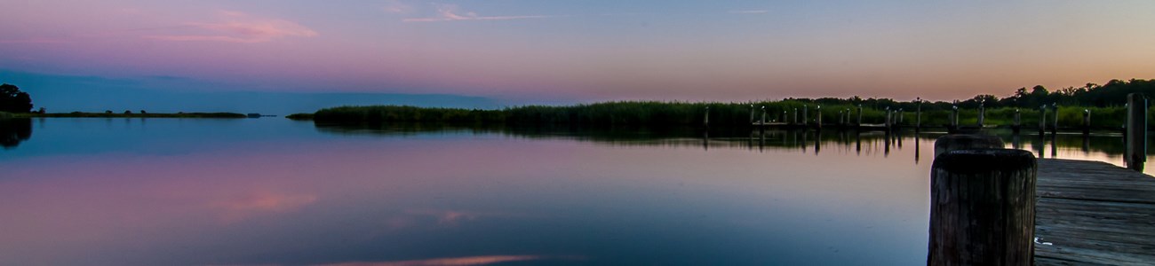 A pink sky fades to the left as the sun begin to rise to the right over water.