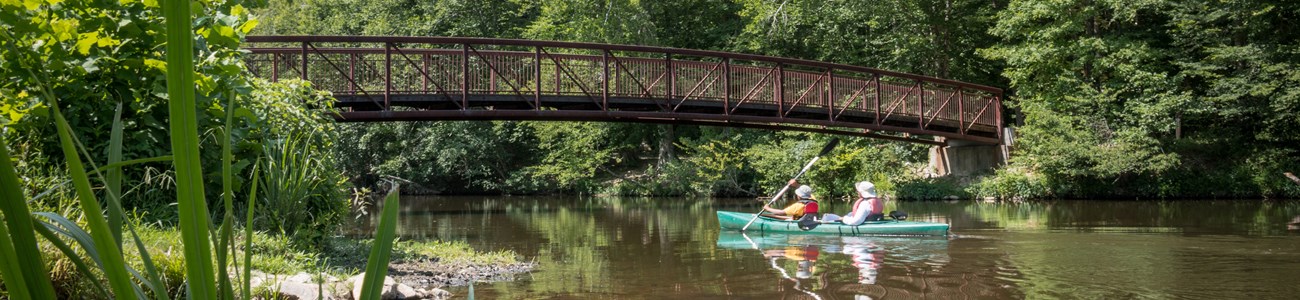 Two people kayak a tandem kayak under a small walking bridge.