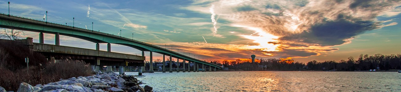 The sun sets behind clouds over a waterway crossed by a concrete bridge.