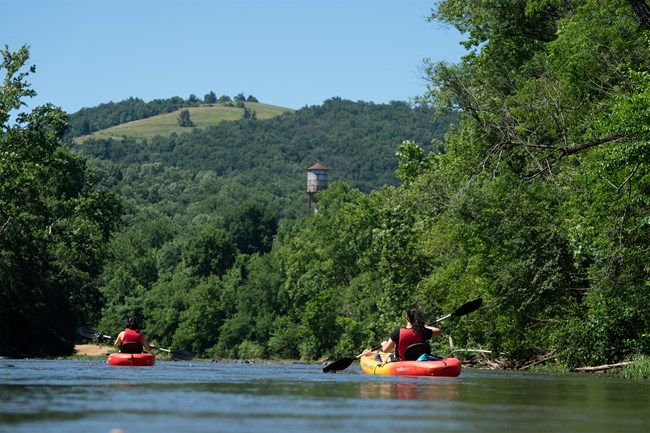 Two kayakers on a river with dense green trees and a distant hill with a water tower.