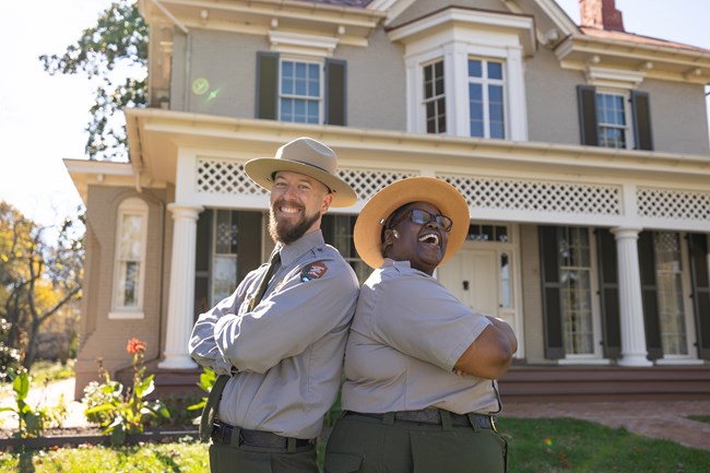 Two rangers pose back to back with arms folded, laughing, in front of the Frederick Douglass House.