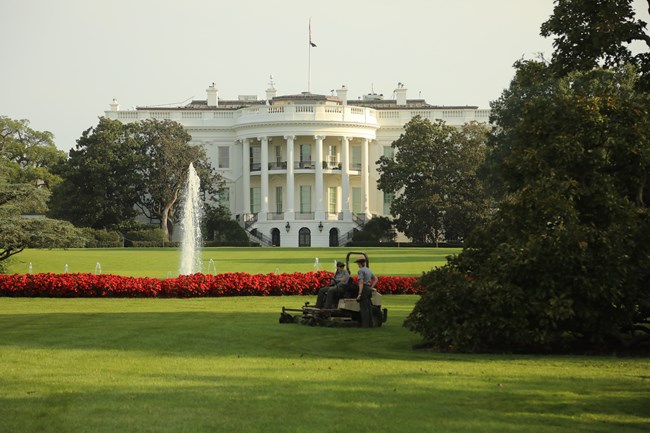 Two National Park employees, one riding on a lawnmower, pause to chat on the White House South Lawn with the White House in the background.