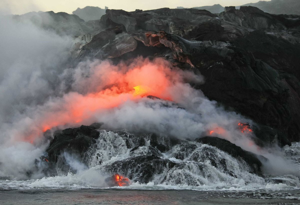 red glowing lava and smoke rise above the ocean and black rocks
