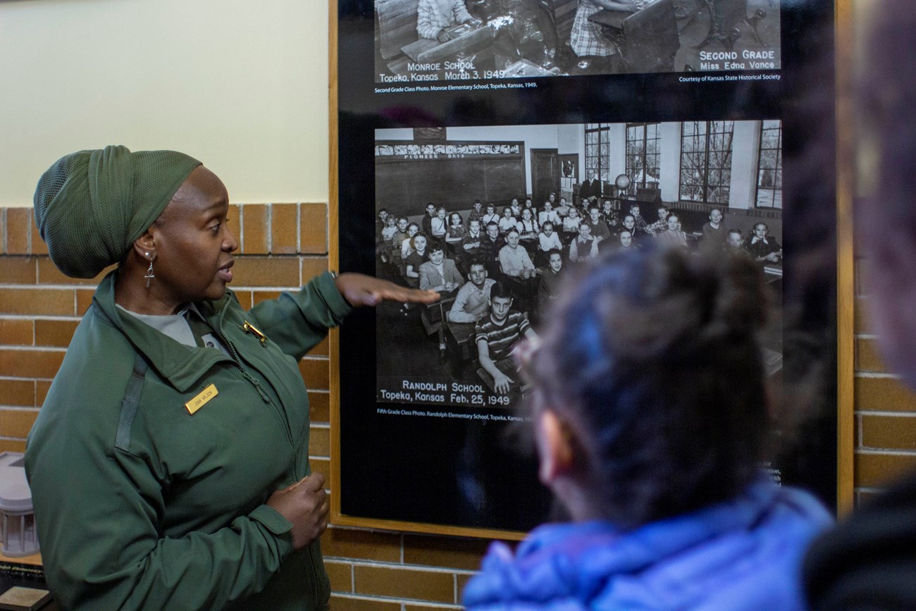 An African American park ranger points to a black and white photo of people inside a building during a program.