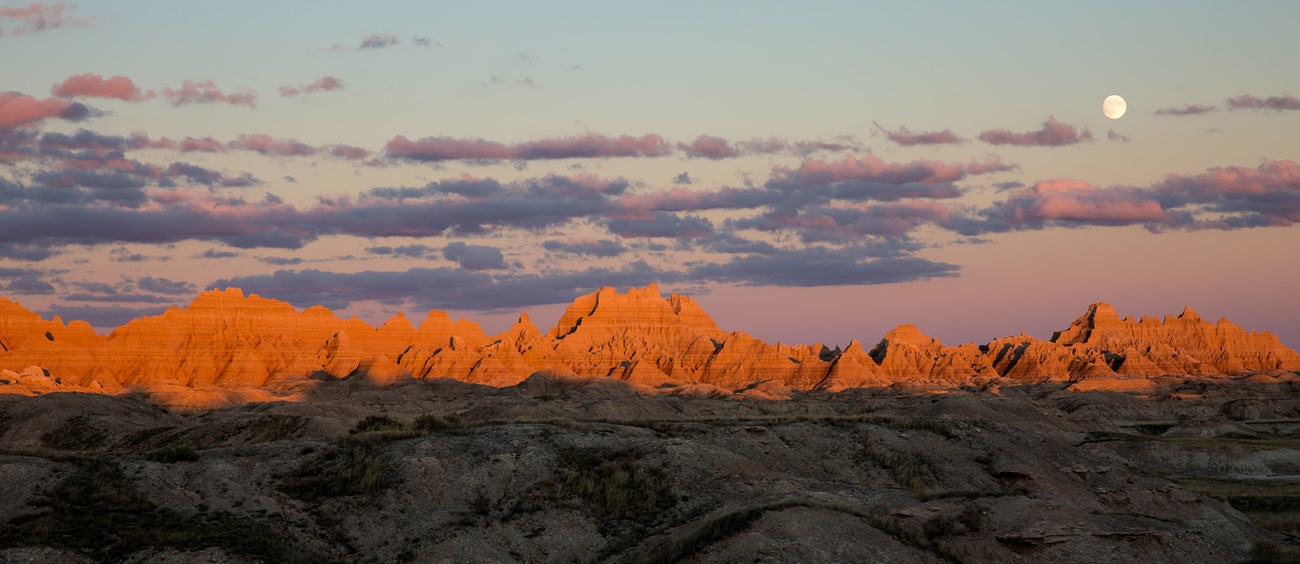 The setting sun illuminates several ridge lines and hills at Badlands National Park. The moon rises in the distance from behind some small clouds.