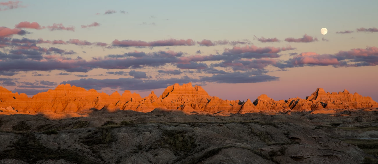 Badlands Moon The setting sun illuminates several ridge lines and hills at Badlands National Park. The moon rises in the distance from behind some small clouds.