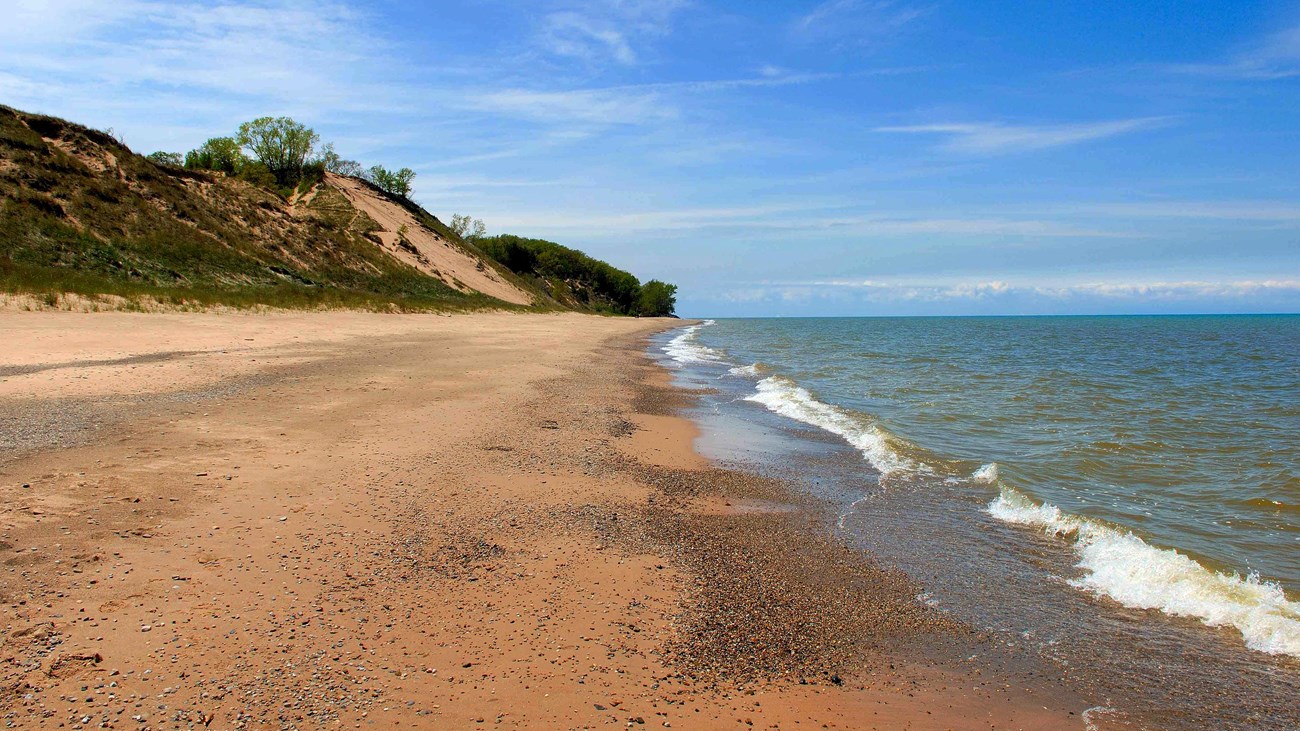 Beach along a large lake with green grassy dunes in the background under a blue sky.