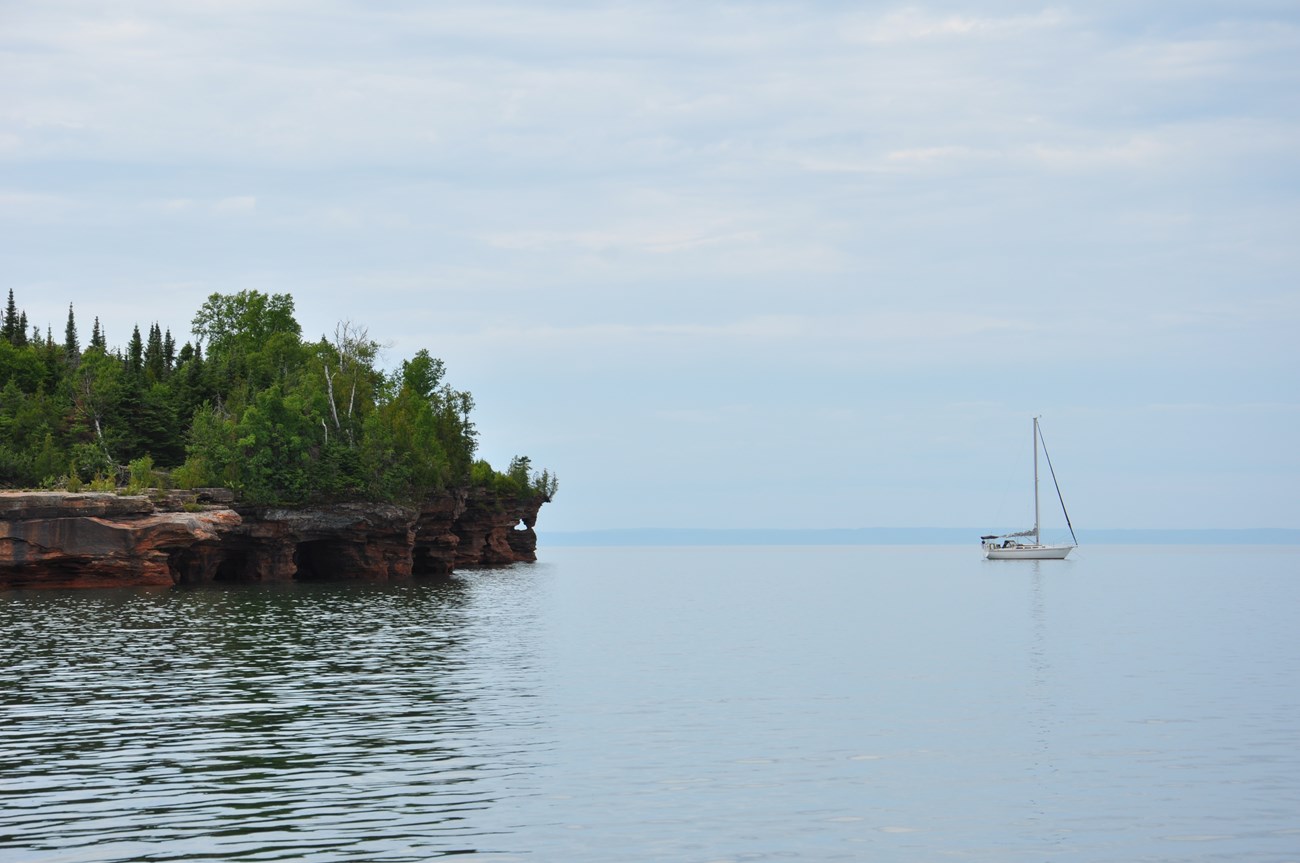 Devil's Island Sailboat A sailboat on calm water near the edge of sandstone sea caves with trees growing on top.