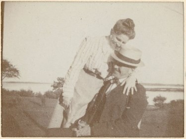 Three-quarter length portrait of a young girl with shoulder-length hair who rests upon a fence in the photographer's studio.  She wears a light-colored ribbon in her hair and her dress is dark-colored.