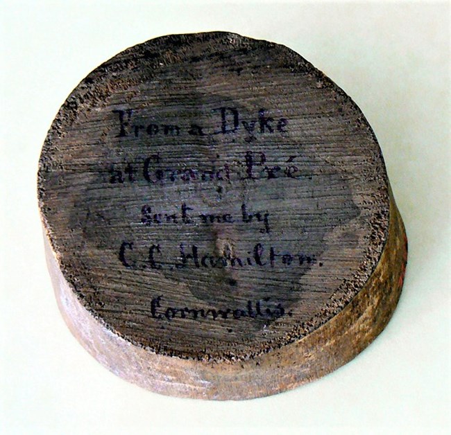 Sliced puck of wood with an inscription in Longfellow handwriting: “From a Dyke / at Grand Pré / Sent me by / C. C. Hamilton. / Cornwallis.”