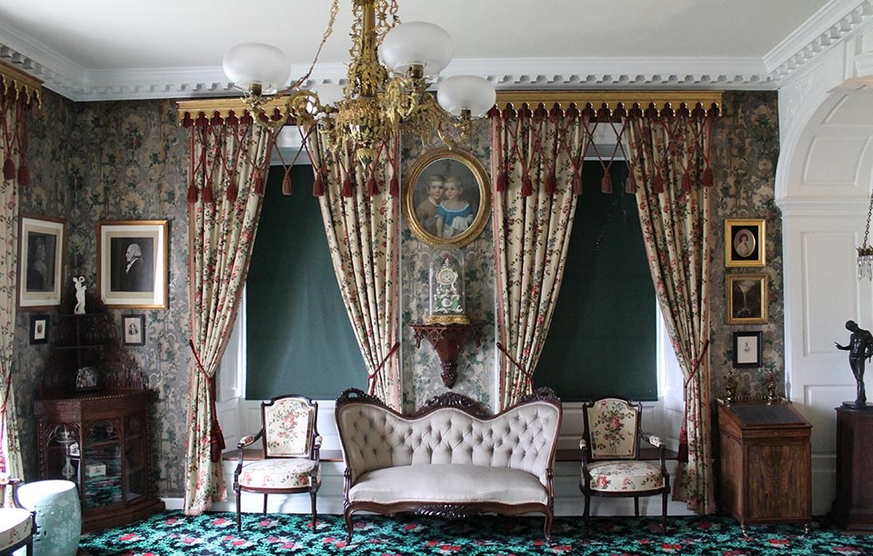 Interior of room with floral wallpaper, drapes, carpet, and upholstery, sofa and two chairs along wall with two windows