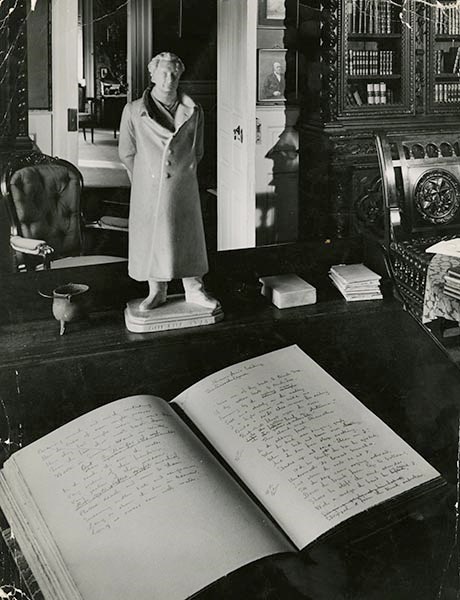 Interior of study with slanted desk in foreground, open handwritten book on it, statue of standing man on ledge at top