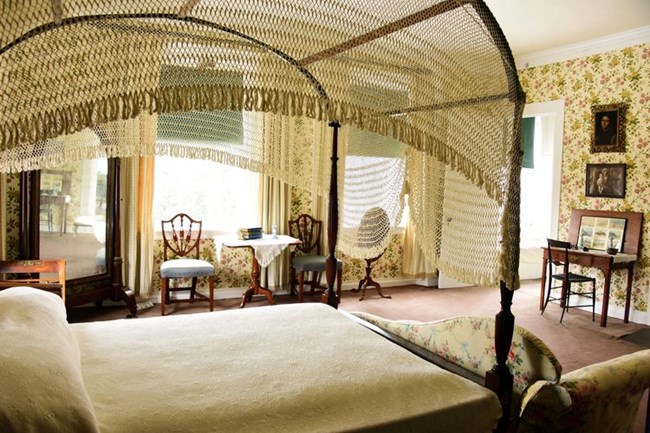 Bedroom with canopy bed in foreground, light floral wallpaper, and tables and chairs against wall with two windows