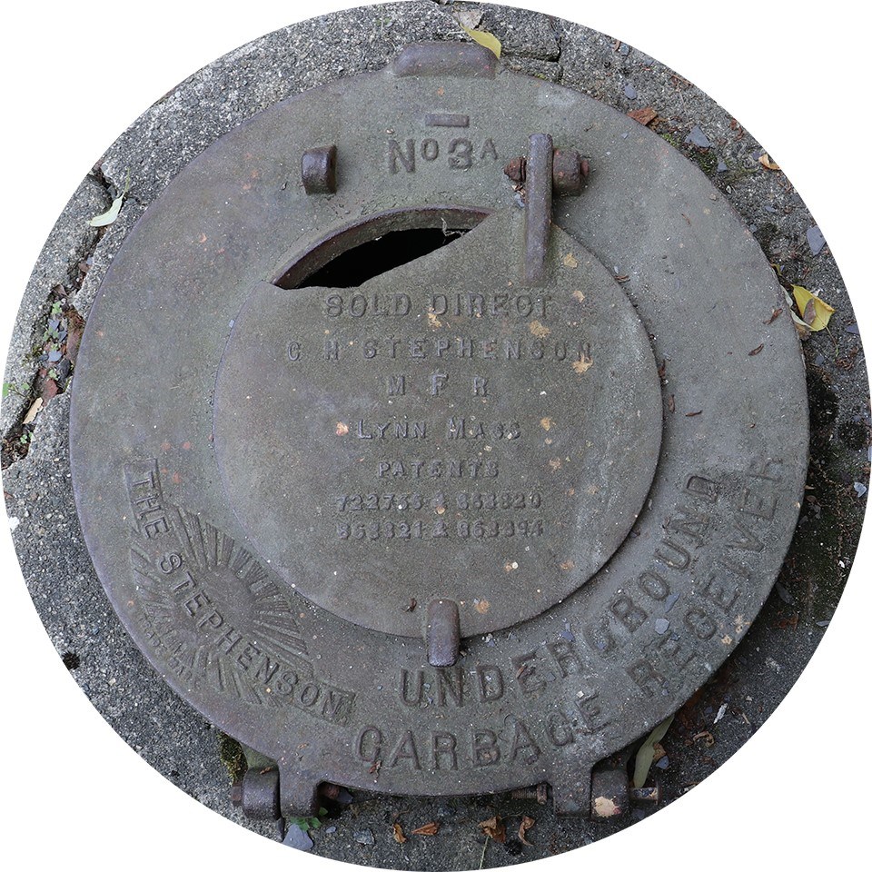 A hinged circular metal lid to an underground garbage receiver with the words Stephenson Underground Garbage Receiver molded on it.