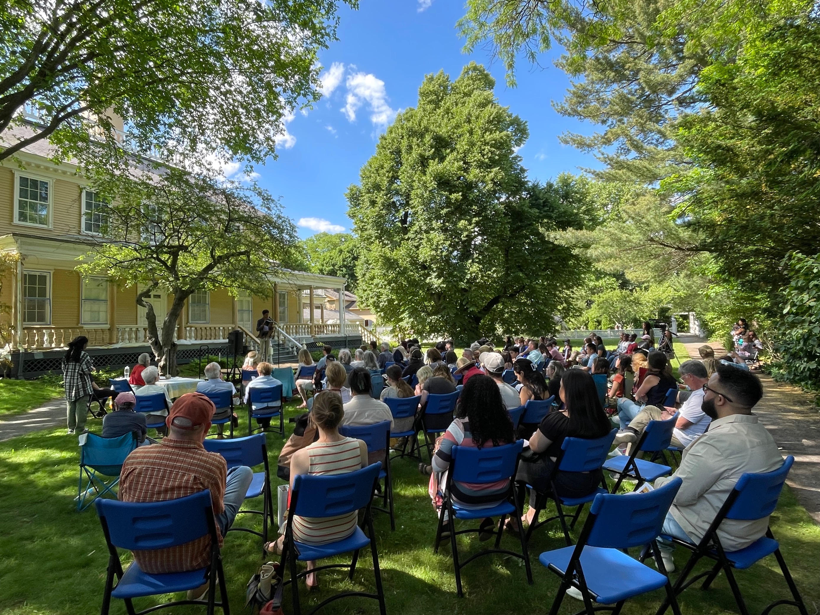 Poet reading on stage with audience outside on the lawn.