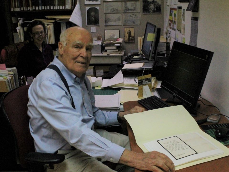 Man sits at computer with open folder of manuscript letters. Woman in glasses sits at desk behind him.