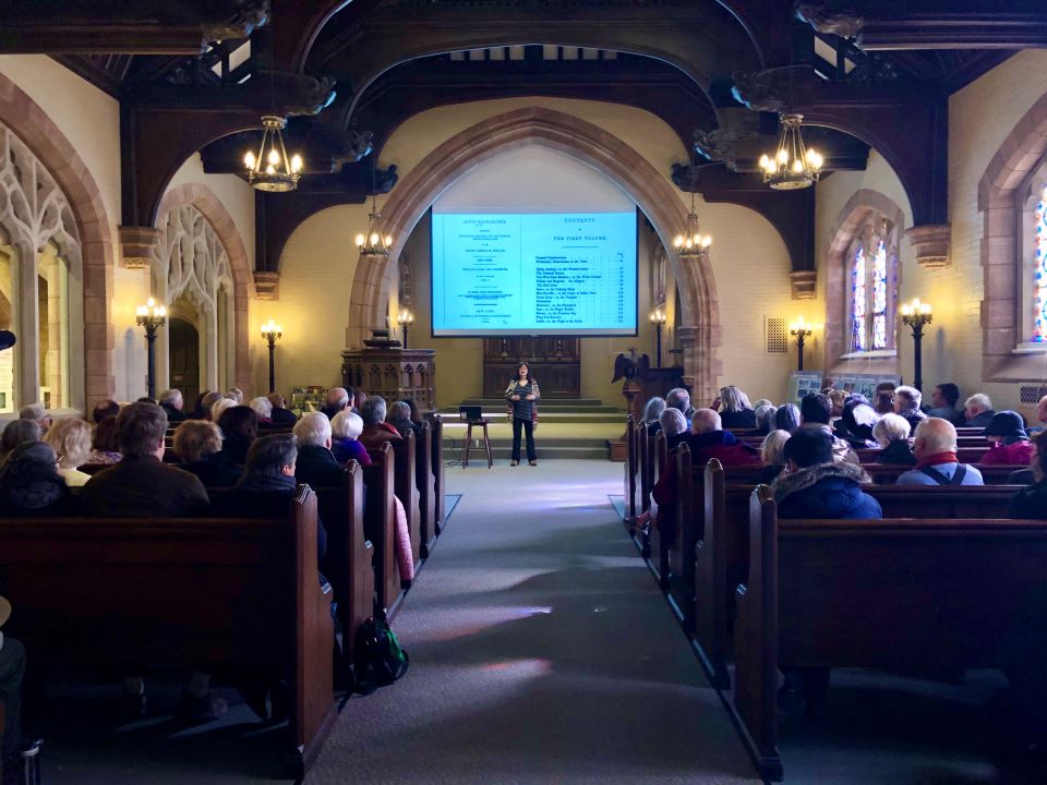 woman stands in front of crowd seated in wooden pews in building with arched stone windows and stained glass