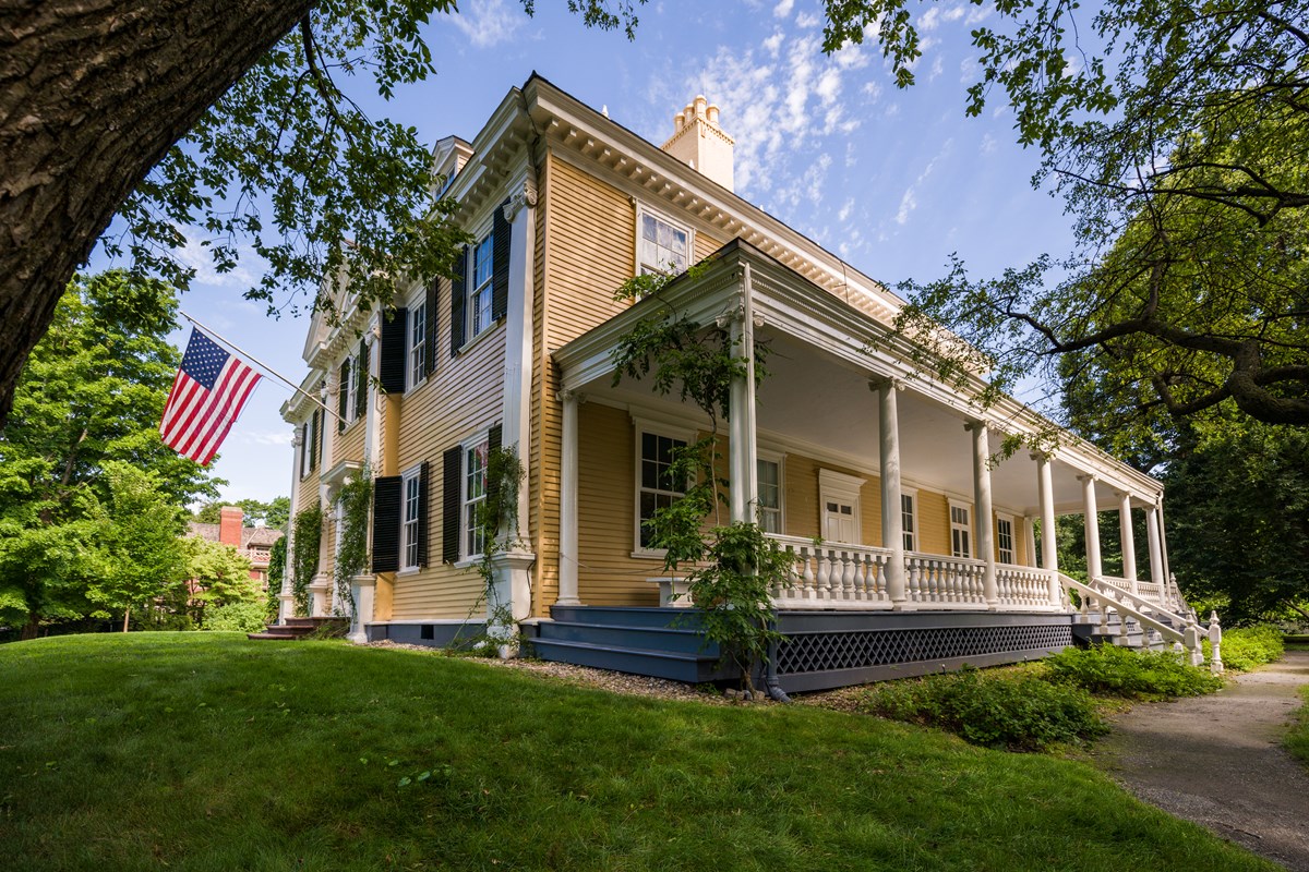 Large yellow mansion with porch along side and US flag flying from front