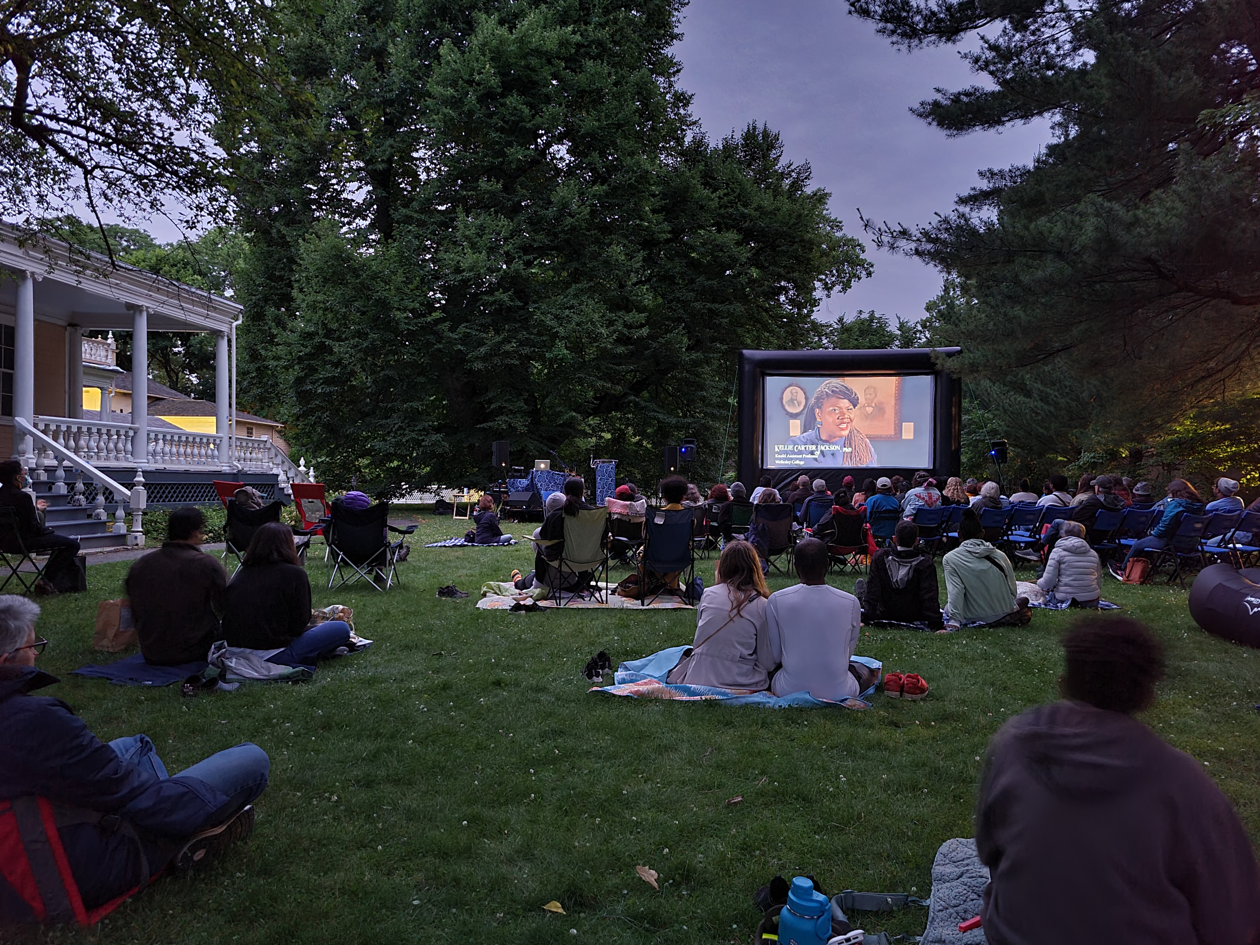 People sit on blankets and lawn chairs outdoors, watching a film at dusk